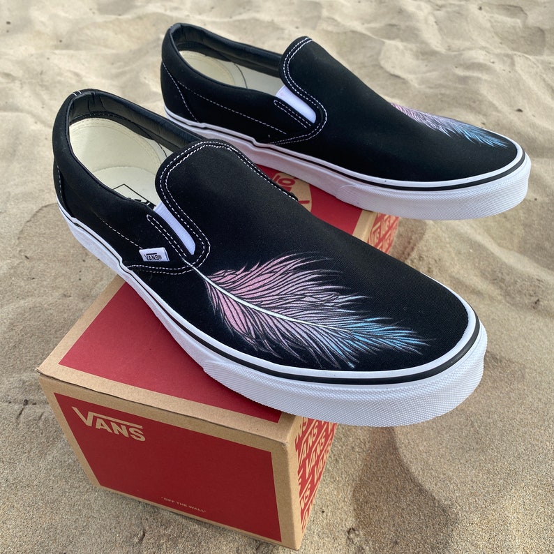 Puede incluir: Un par de zapatos Vans negros de lona, sin cordones, con un dise&ntilde;o pintado de una pluma rosa, azul y blanca en la punta de cada zapato. Los zapatos est&aacute;n sobre una caja de cart&oacute;n marr&oacute;n con el logotipo de Vans impreso en el lateral.
