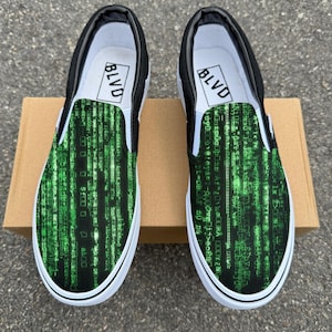 May include: Pair of slip-on trainers with a black and white design. The top of the shoe features a green digital rain pattern. The inner sole has the word "BLVD" printed in a white rectangle. The shoes are on a brown cardboard box.