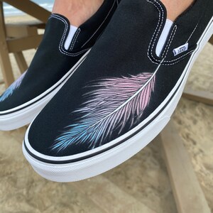 Puede incluir: Zapatillas de lona negras de deslizamiento con suela blanca y un dise&ntilde;o de pluma rosa, azul y blanca pintado en el lateral. Las zapatillas tienen la palabra "VANS" impresa en el lateral.