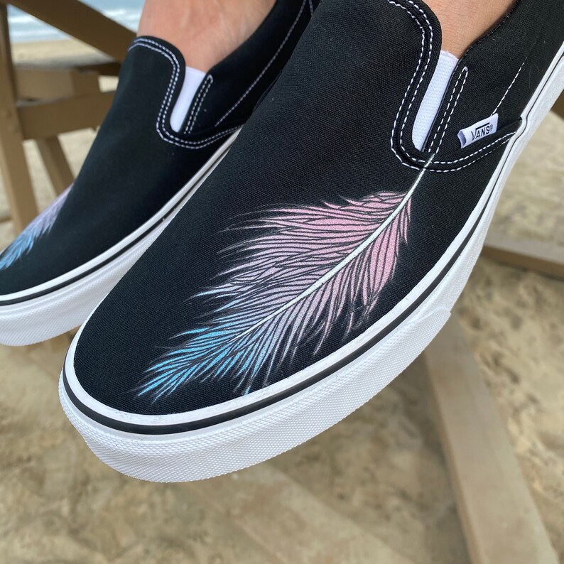 Puede incluir: Zapatillas de lona negras de deslizamiento con suela blanca y un dise&ntilde;o de pluma rosa, azul y blanca pintado en el lateral. Las zapatillas tienen la palabra "VANS" impresa en el lateral.