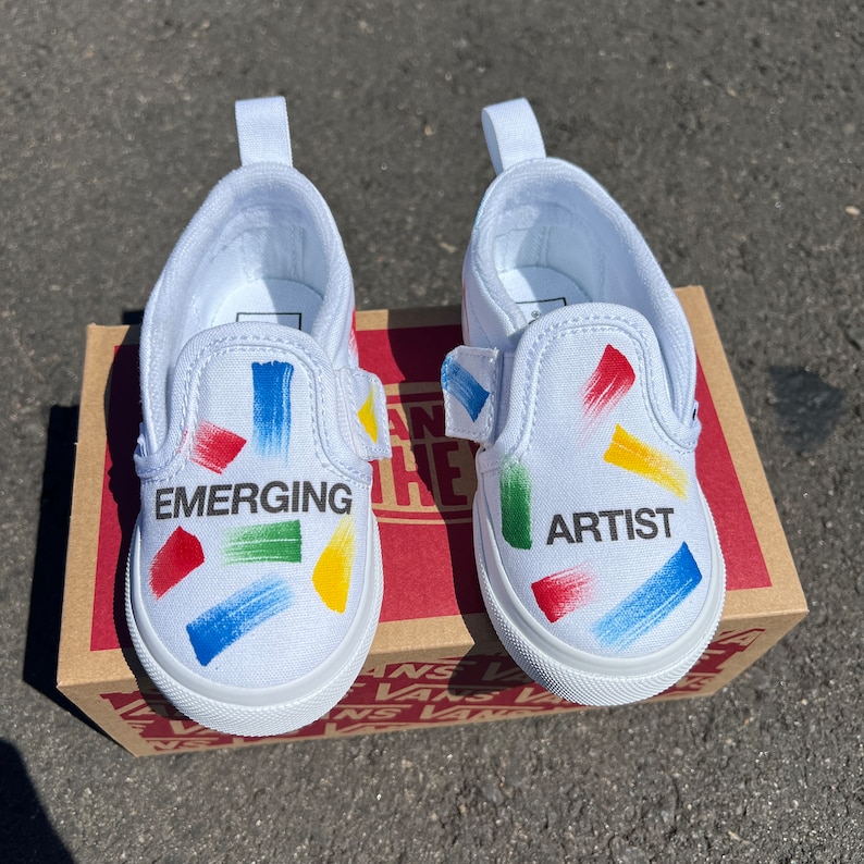 May include: A pair of white slip-on Vans shoes with colourful paint splatter designs. One shoe says "Emerging" and the other says "Artist".