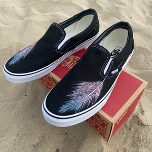 Puede incluir: Un par de zapatos de lona negros de deslizamiento con un dise&ntilde;o de pluma blanca pintado en el lateral. La pluma es rosa y azul.