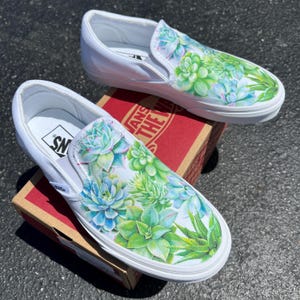 May include: A pair of white canvas slip-on shoes with a hand-painted design of succulents in shades of green and blue.