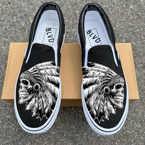 May include: Pair of black slip-on shoes with a white sole. Each shoe features a detailed black and white graphic of a skull wearing a feathered headdress. The inside of the shoe has the word "BLVD".
