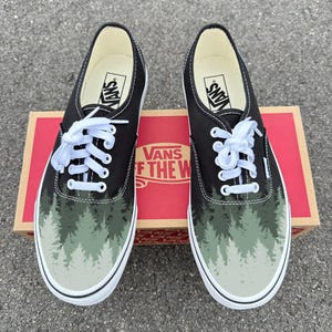 Könnte beinhalten: Paar schwarze Vans-Sneaker mit einem Waldmotiv auf der Spitze. Die Schuhe haben weiße Schnürsenkel, eine weiße Sohle und das Vans-Logo auf der Zunge. Die Schuhe werden auf einer roten und braunen Vans-Box präsentiert.
