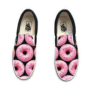 Könnte beinhalten: Paar Slip-On-Sneaker mit schwarzem Hintergrund und einem Muster aus rosa Donuts mit bunten Streuseln. Die Schuhe haben schwarze Verzierungen, weiße Sohlen und das Vans-Logo auf der Innensohle.
