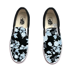 Könnte beinhalten: Paar schwarze Slip-On-Sneaker mit einem hellblauen Blumenmuster. Die Schuhe haben eine weiße Sohle und schwarze Zierleisten. Der Markenname "Vans" ist auf dem Innenetikett sichtbar.