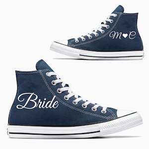 May include: A pair of navy blue high-top sneakers with white laces. One shoe says "Bride" in white cursive lettering and the other says "M♥E" in white cursive lettering.