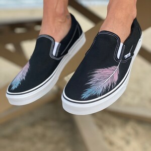 Puede incluir: Zapatillas de lona negras de deslizamiento con un dise&ntilde;o de pluma blanca en el lateral. La pluma es rosa, azul y blanca.