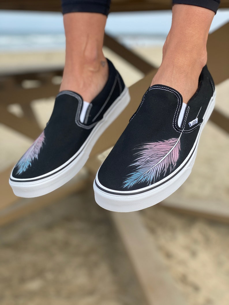 Puede incluir: Zapatillas de lona negras de deslizamiento con un dise&ntilde;o de pluma blanca en el lateral. La pluma es rosa, azul y blanca.