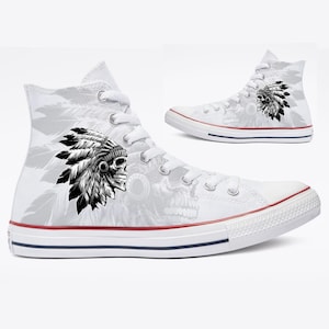 May include: White high-top sneakers with a black and white Native American headdress skull design. The shoes have a red and blue stripe on the sole and white laces. The design is repeated on the side of the shoe.