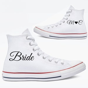 May include: Two white high-top sneakers. One sneaker has the word "Bride" in black script. The other sneaker has the initials "M" and "C" with a heart symbol in between. The sneakers have red and blue stripes on the soles.