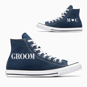 May include: Two navy blue high-top sneakers. One sneaker has the word "GROOM" in white lettering. The other sneaker has the initials "M C" with a heart symbol. Both sneakers have white laces, soles, and toe caps.