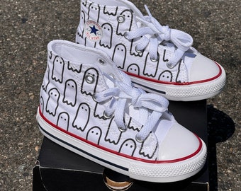 May include: A pair of white high-top Converse sneakers with a black ghost pattern. The shoes are on a black box with the Converse logo.