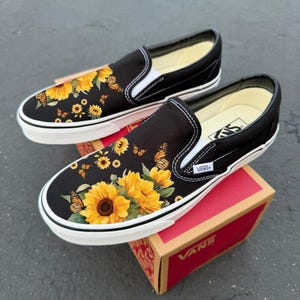 Könnte beinhalten: Ein Paar schwarze Slip-on-Sneaker mit einem Druckmuster aus gelben Sonnenblumen und orangefarbenen Monarchfaltern auf der Vorderkappe. Die Schuhe haben weiße Gummisohlen, weiße elastische Seiteneinsätze und ein kleines weißes Vans-Markenetikett.
