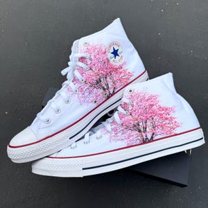 以下が含まれることがあります： ピンクの桜の木のデザインが施された白いハイカットのコンバーススニーカー。スニーカーには、青い星形のロゴと「Converse All Star」の文字が書かれています。