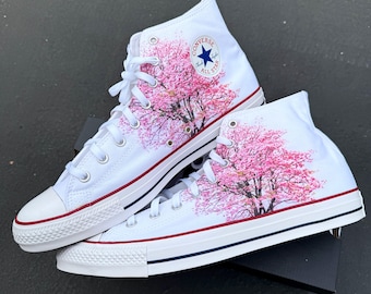 May include: White high-top Converse sneakers with a pink cherry blossom tree design. The sneakers feature a blue star logo with the text "Converse All Star".