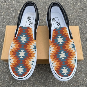 May include: Pair of slip-on shoes with a geometric pattern in shades of orange, blue, and cream. The shoes have black accents, white soles, and the word "BLVD" printed on the inner lining.
