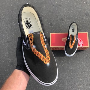 Może przedstawiać: Czarne slip-ony Vans z wzorem złotego łańcucha. Buty mają białą podeszwę, a logo Vans jest widoczne na tylnej części buta.