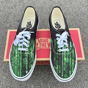 Może przedstawiać: Para czarnych i zielonych butów sportowych Vans z wzorem w stylu matrycy. Buty mają białe sznurowadła i podeszwy oraz czarną cholewkę. Logo Vans jest widoczne na języku i wkładce. Buty są prezentowane na pudełku Vans.