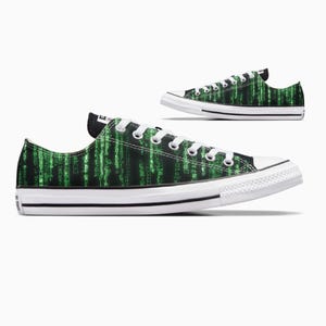 May include: Low-top canvas trainers featuring a black and green digital rain design. The shoes have white laces, a white rubber toe cap, and a black heel patch. Another trainer is visible in the background.