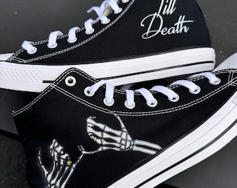 Op de afbeelding: Een paar zwarte high-top canvas sneakers met witte veters. Eén schoen heeft de tekst "Till Death" in witte letters op de zijkant. De andere schoen heeft een afbeelding van twee skelethandjes die elkaar vasthouden met gouden ringen aan hun vingers.