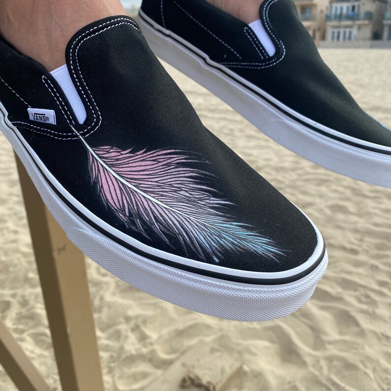 Puede incluir: Zapatillas Vans negras de slip-on con suela blanca y un dise&ntilde;o de pluma rosa y azul en la parte superior del zapato.