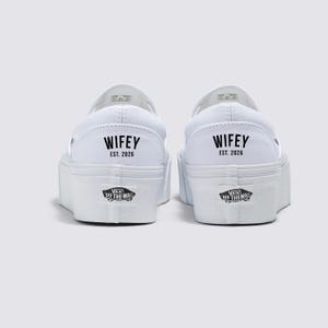Könnte beinhalten: Zwei weiße Slip-On-Sneaker mit Plateau. Auf der Ferse jedes Schuhs steht in schwarzer Schrift "WIFEY EST. 2026". Das Vans-Logo ist auf der Rückseite jeder Sohle zu sehen.
