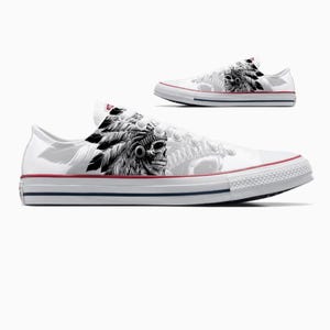 May include: White canvas sneakers with a black and white Native American headdress and skull design. The shoes have a red stripe along the top of the sole and a blue stripe below. The design is on the side of the shoe.