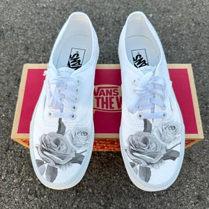 Może przedstawiać: Para białych trampek Vans z szarym wzorem róż. Buty leżą na pudełku po butach Vans. Logo Vans jest widoczne na języku i wkładce.