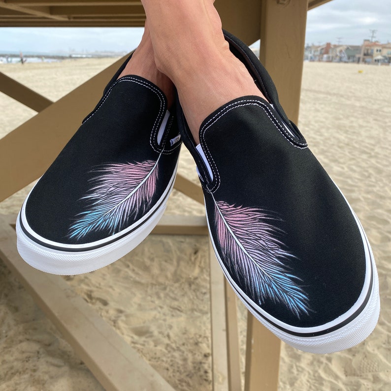 Puede incluir: Un par de zapatos de lona negros de deslizamiento con un dise&ntilde;o de pluma blanca. La pluma es rosa y azul, asemej&aacute;ndose a una bandera del orgullo transg&eacute;nero.