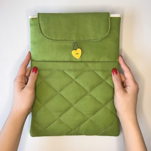 May include: A green quilted laptop sleeve with a flap closure. The sleeve has a yellow heart-shaped button and a white interior lining. The hands holding the sleeve have red nail polish.