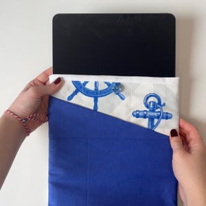 Puede incluir: Funda para tablet de temática náutica en azul y blanco. Presenta un diseño de ancla y timón azul sobre fondo blanco, con una base azul lisa. Diseñada para proteger una tablet.
