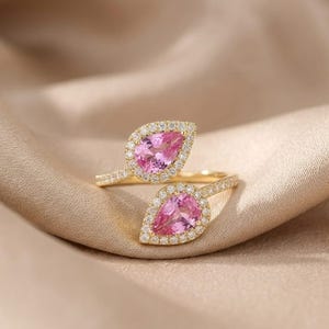 May include: A gold bypass ring featuring two pear-shaped pink gemstones, each surrounded by a halo of small white round-cut stones. The band is thin and metallic, with the two teardrop-shaped settings positioned to face each other at the centre.