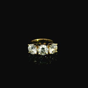 May include: A gold ring with three large, clear gemstones set in a row. The ring is set against a black background, highlighting the sparkle of the stones. The ring's band is a simple, classic design.