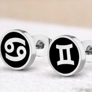 Puede incluir: Un par de pendientes de acero inoxidable. Cada pendiente presenta un círculo negro con un símbolo zodiacal blanco. Uno muestra el símbolo de Cáncer y el otro el de Géminis. Los pendientes están sobre una superficie blanca.