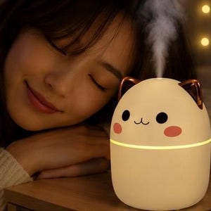Op de afbeelding: Een beige katvormige etherische olie diffuser met een lachend gezicht, zwarte ogen en roze wangen. De diffuser heeft koperkleurige oren en geeft een stroom stoom af. Een vrouw rust haar hoofd naast de diffuser.