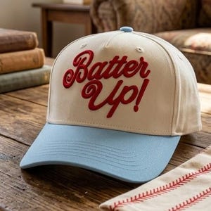 Puede incluir: Una gorra de béisbol con corona de color beige claro y visera azul claro. El texto "Batter Up!" está bordado en rojo en la parte delantera. Una servilleta crema doblada con costuras rojas y una taza de cerámica también están sobre la mesa de madera.