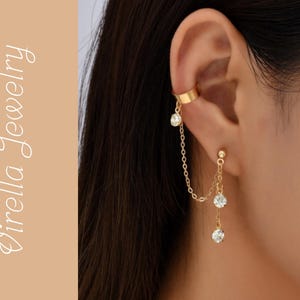 Puede incluir: Puño de oreja y pendientes colgantes dorados con gemas redondas y transparentes. El puño de oreja se envuelve alrededor de la parte superior de la oreja, conectado por una delicada cadena a los pendientes. El texto "Virella Jewelry" está a la izquierda.