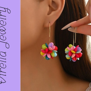 Op de afbeelding: Goudkleurige oorhangers met kleurrijke, geclusterde bloemmotieven. De oorbellen hebben een mix van roze, rode, gele, blauwe en paarse bloemblaadjes, wat een speels accessoire creëert. De tekst "Virella Jewelry" staat links.