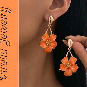 Könnte beinhalten: Goldfarbene Ohrringe mit orangefarbenen Blumenanhängern. Die Ohrringe haben einen Steckerverschluss und einen tropfenförmigen Akzent. Das Blumendesign hat eine detaillierte Mitte und ein glänzendes Finish. Der Text "Virella Jewelry" befindet sich auf der linken Seite.