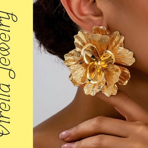 Peut inclure: Boucles d'oreilles florales dorées. Les boucles d'oreilles sont grandes, avec des textures de pétales détaillées. L'image comprend également le texte "Virella Jewelry" en orientation verticale sur fond jaune.