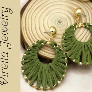 May include: Olive green raffia earrings with gold-colored accents. The earrings are circular with a woven design and a gold post. The earrings are displayed on a wooden slice. The text "Virella Jewelry" is on the left side.
