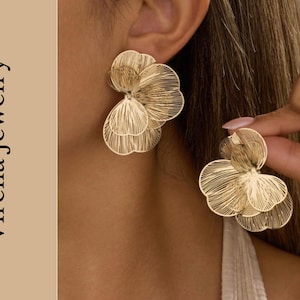 May include: Gold-coloured floral earrings with a layered, openwork design. The earrings are in the shape of a flower with multiple petals. The text "Virella Jewelry" is on the left side of the image.
