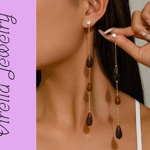 May include: Gold-toned, long drop earrings featuring teardrop-shaped brown beads and small clear crystals. The earrings are suspended from the ear. The image includes the text "Virella Jewelry" in a script font on a lavender background.