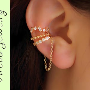 Puede incluir: Un conjunto de pendientes tipo ear cuff dorados con tres bandas apiladas con piedras transparentes y una delicada cadena colgante. La banda superior tiene piedras redondas y baguette, la central es de cuentas y la inferior tiene piedras redondas.
