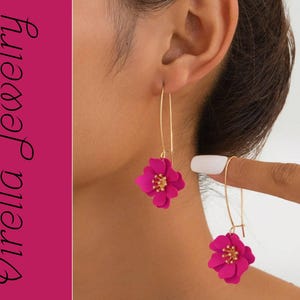 Op de afbeelding: Goudkleurige oorhangers met fuchsia bloemhangers en goudkleurige centra. De oorbellen worden getoond aan het oor van een persoon. De afbeelding bevat ook de tekst "Virella Jewelry" in een cursief lettertype.