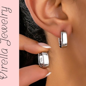 Puede incluir: Pendientes de aro plateados brillantes con diseño rectangular. Una mano con una uña manicurada sostiene un pendiente. Los pendientes están marcados con "925". El texto "Virella Jewelry" está en el lado izquierdo de la imagen.
