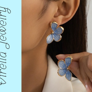 May include: A pair of floral earrings with a light blue enamel finish and gold-colored trim. The earrings are in the shape of a flower with three petals. The image also includes the text "Virella Jewelry" in a script font.
