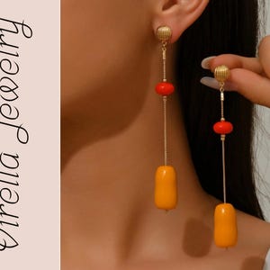 Puede incluir: Pendientes colgantes dorados con detalles rojos y amarillos. Los pendientes presentan un pequeño perno dorado texturizado, una cuenta roja y una larga cuenta oblonga amarilla. Los pendientes son parte de la colección Virella Jewelry.